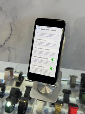 A black smartphone displayed on a metallic charging stand, showing a battery optimization settings screen. The stand is placed on a white marble-patterned surface with blurred watch displays in the background.