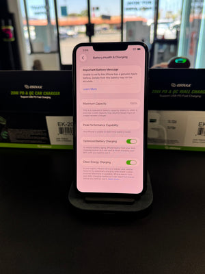 A black smartphone lying on a dark charging dock or stand, with its screen lit and showing battery settings. Behind it are product boxes, one with visible GESSER branding, against a dark indoor background.