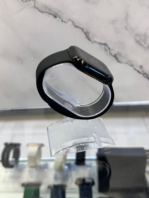 A black smartwatch with a matte black band displayed on a clear acrylic stand, set against a marble-patterned background.