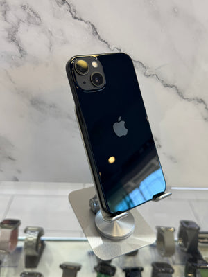A black Apple iPhone 11 standing upright on a silver display stand, showing the back of the phone with its dual camera system and Apple logo. The background features a white marble pattern wall and a glass counter displaying other devices.