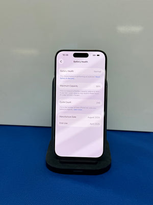 A black smartphone displayed on a black charging stand against a light gray wall, with a blue surface underneath. The phone screen shows the Battery Health settings page.