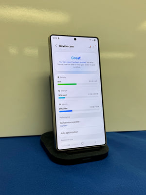 A smartphone displayed upright on a black charging stand against a pale yellow wall, with a blue surface underneath. The phone screen shows a device care settings page.