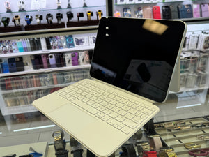 A cream-colored iPad with a matching keyboard cover and a black screen, displayed on a stand inside a retail store with shelves of accessories in the background.