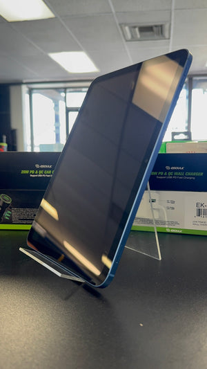 A blue tablet standing upright on a clear acrylic display stand on a dark tabletop, with product boxes in the background and a real indoor office environment visible.