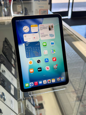 A black Apple iPad tablet on a clear display stand inside a retail store, showing the home screen with various app icons.