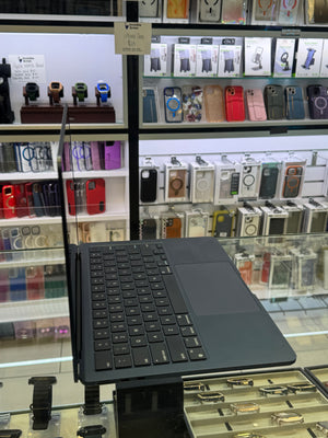 A black Microsoft Surface Laptop with attached keyboard and trackpad displayed on a stand inside a retail store, with shelves of phone cases and accessories in the background.