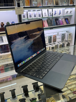 A dark gray Apple MacBook laptop with an open screen displaying a mountain landscape wallpaper, set on a glass display counter inside a retail store with shelves of accessories in the background.