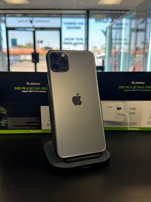 A silver Apple iPhone Pro standing upright on a charging dock on a dark tabletop, with retail packaging and a blurred storefront visible in the background.