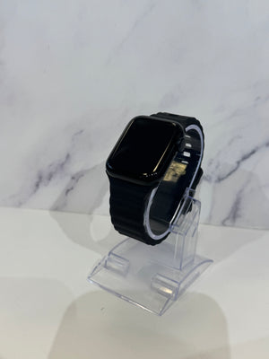 A black smartwatch with a rectangular black screen and black band, displayed on a clear acrylic stand against a white marble-patterned background.