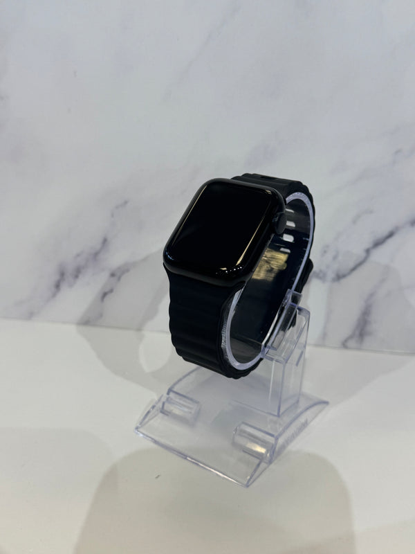 A black smartwatch with a rectangular black screen and black band, displayed on a clear acrylic stand against a white marble-patterned background.