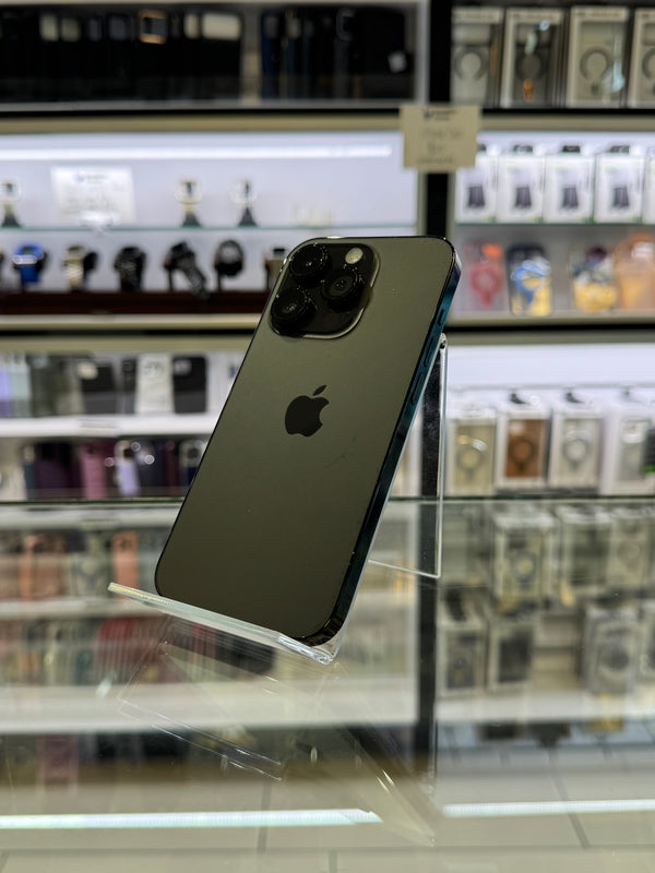 A dark gray Apple iPhone Pro shown from the back, suspended by a clear plastic sleeve, with a blurred retail store background featuring display cases.