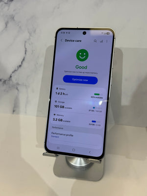 A smartphone displayed upright on a metal stand, showing the Google 'Device care' screen with battery, storage, and memory details. The phone is photographed against a light marble-patterned background.