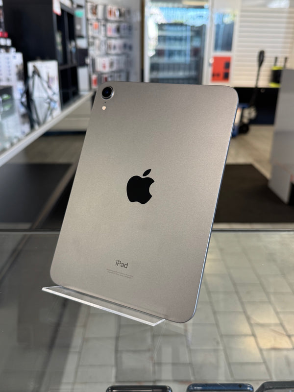 A silver Apple iPad shown from the back, resting on a clear display stand inside a retail store. The Apple logo is clearly visible in the center.