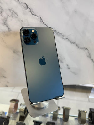 An Apple iPhone 12 Pro in blue-gray color, shown from the back on a silver metal display stand against a white marble-patterned background.