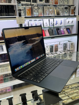 A dark gray Apple MacBook laptop with an open screen displaying a scenic wallpaper, set on a glass display counter inside a retail store with shelves of phone accessories in the background.