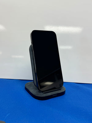 A black smartphone with a glossy screen stands upright on a dark gray charging dock or stand against a white background, with a blue surface visible at the bottom.