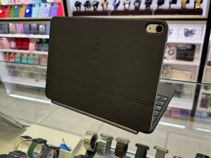 A black Apple iPad with a magnetic keyboard case, shown from the back in a retail store setting with blurred watch and accessory displays in the background.