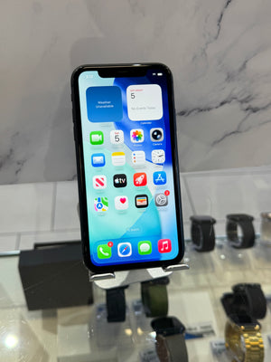 A black smartphone with a lit home screen displaying various app icons, standing on a small display stand in a retail setting with marble-patterned walls and other watches or accessories blurred in the background.