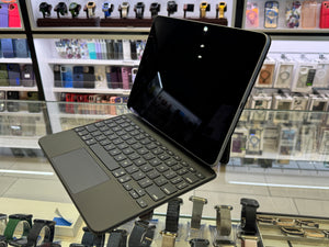A black tablet in a matching keyboard case with a touchpad, displayed on a glass retail counter with watch and phone accessories in the background.