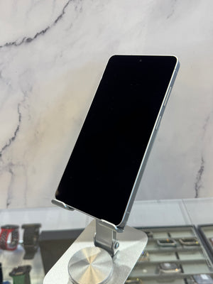 A black smartphone with a silver edge stands on a metallic display stand in front of a light marble-patterned wall.