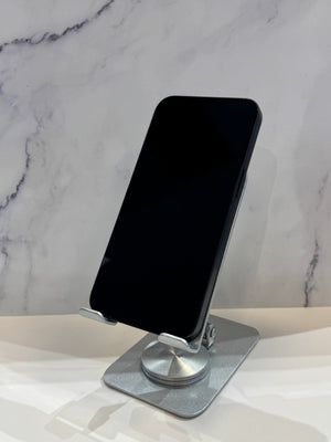 A silver metal phone stand holding a black smartphone, set on a white surface against a gray marble-patterned background.