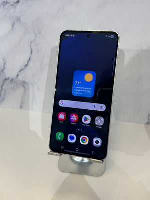 A Samsung smartphone with a black screen displaying the home interface, resting on a silver metal display stand against a white marble-patterned background.