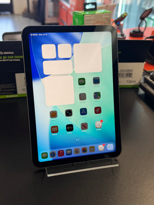 A black iPad Pro tablet with a colorful home screen, displayed on a clear acrylic stand on a dark tabletop. The background shows a busy retail or office environment with boxes and equipment.