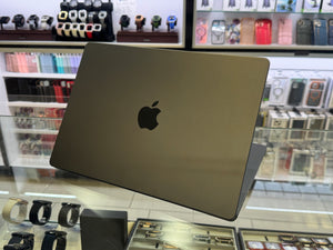 A closed Apple MacBook laptop in dark gray aluminum sits on a glass display counter inside a retail store, with watch and accessory displays blurred in the background.