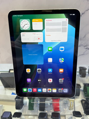 A black Apple iPad tablet with an active home screen, displayed upright on a retail counter with other watches in the background.
