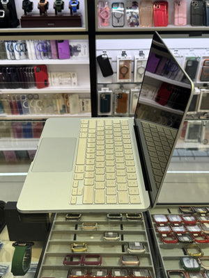 A silver aluminum laptop with a cream-colored keyboard cover and trackpad, shown open on a display stand inside a retail store with shelves of phone cases and watches in the background.