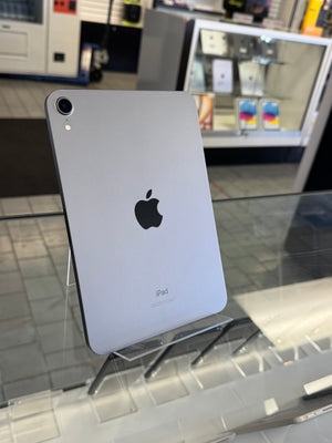 A silver Apple iPad shown from the back, standing on a clear display stand inside a retail store. The Apple logo is visible in the center, with a glass display counter and other store fixtures blurred in the background.