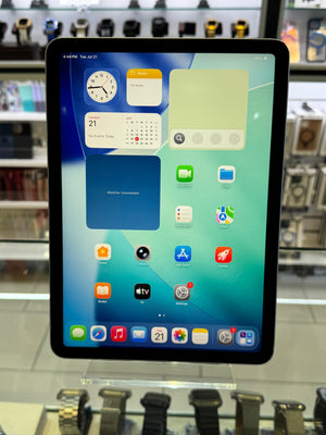 An iPad displaying its home screen with various app icons, set against a blurred retail background showing other electronic accessories.