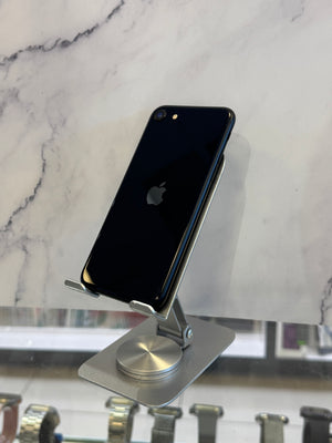 A black Apple iPhone with a glossy finish, shown from the back, resting on a silver metal display stand against a white marble-patterned background.