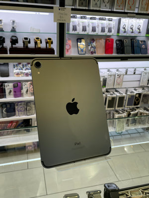 A silver Apple iPad shown from the back, resting on a glass display counter inside a retail store with other electronics and accessories visible in the background.