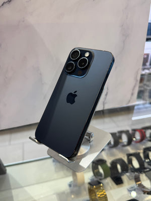 A dark blue-gray Apple iPhone 12 Pro Max displayed on a silver stand in a retail setting, with a marble-patterned wall and watch displays in the background.