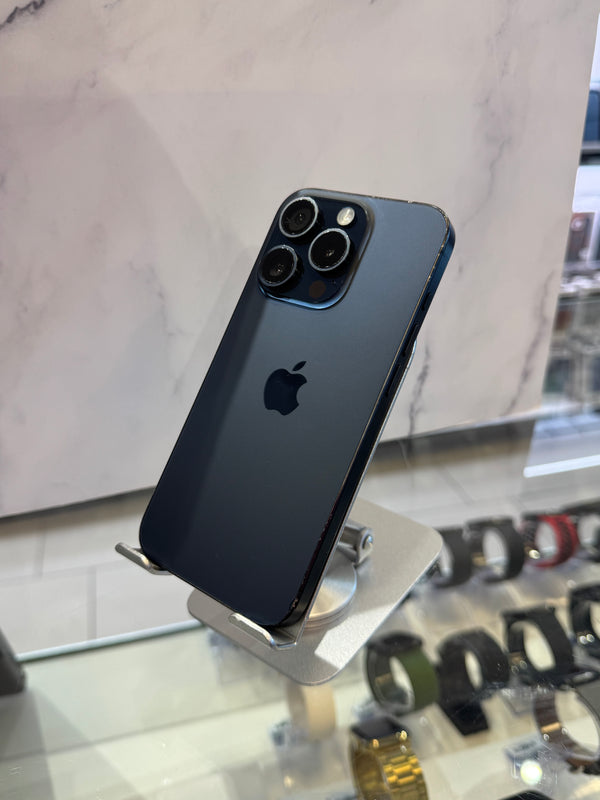 A dark blue-gray Apple iPhone 12 Pro Max displayed on a silver stand in a retail setting, with a marble-patterned wall and watch displays in the background.