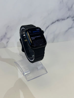 A black smartwatch with a rectangular digital display stands on a clear acrylic display stand against a white marble-patterned background.