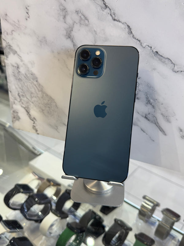A dark blue-gray Apple iPhone 12 Pro shown from the back, resting on a white marble-patterned surface with a watch display case visible in the background.