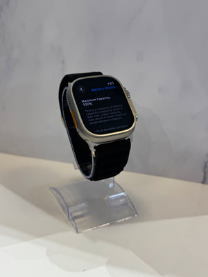 A smartwatch with a black strap and silver-toned case, displayed on a clear acrylic stand against a light gray marble-patterned background.