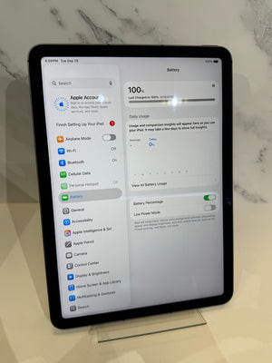 An iPad displaying the Apple Accurate battery settings screen, standing on a small clear display stand against a light marble-patterned background.