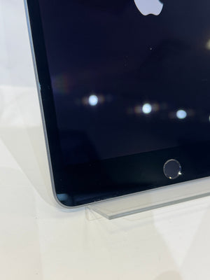 A close-up shot of a black Apple iPad showing its front bezel, home button, and part of the Apple logo on the screen, resting on a clear acrylic display stand.