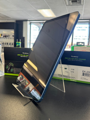 A blue and black Apple iPad standing on a clear acrylic display stand on a dark countertop, with its retail packaging boxes visible in the background inside a store.