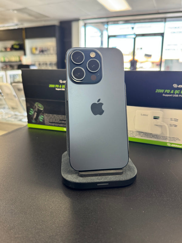 A dark gray Apple iPhone Pro displayed upright on a charging stand on a dark countertop, with Apple product boxes visible in the blurred background of a retail store.
