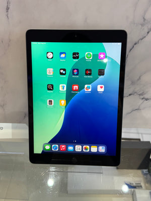 A black tablet, likely an iPad, standing upright on a clear display stand. The screen is on and displaying its home screen with various colorful app icons against a green and blue wallpaper.