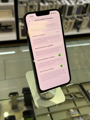 A smartphone displaying the Battery Health & Charging settings screen, photographed standing on a display stand inside a retail store with watch cases blurred in the background.