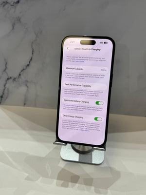 A black smartphone displayed upright on a silver stand, showing the Battery Health & Charging settings screen. The phone is photographed on a glossy white surface against a light gray marble-patterned background.