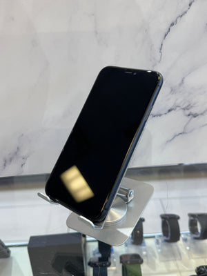 A black smartphone with a silver edge stands on a small metallic display stand in a retail setting, with a marble-patterned wall behind it and other devices partially visible below.
