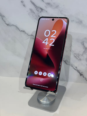 A smartphone with a black front and a dark red or purple side frame stands on a small metallic display stand against a white marble-patterned background.