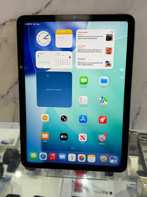An iPad with a black bezel displaying the home screen, standing on a glass display stand in a retail or showroom setting.