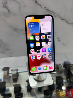 A black iPhone with its home screen on, displayed on a silver stand in a retail setting with a marble-patterned background and other watches blurred in the foreground.
